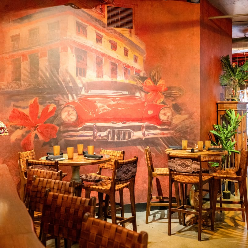Havana 151 restaurant interior with a mural of a red vintage car and buildings, and woven leather chairs.