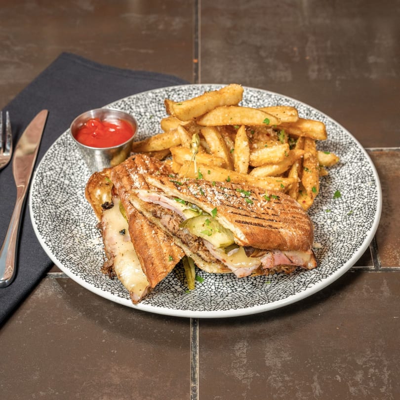 Cuban sandwich with fries and ketchup on a patterned plate, with a fork and knife.