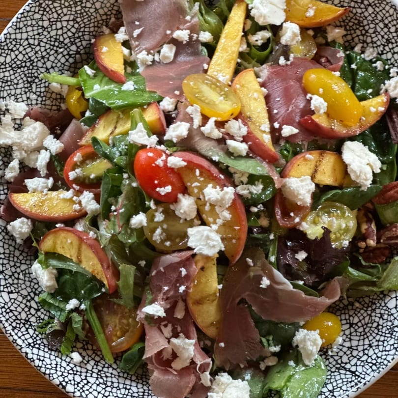 A vibrant salad with prosciutto, grilled peaches, tomatoes, and feta cheese in a patterned bowl.