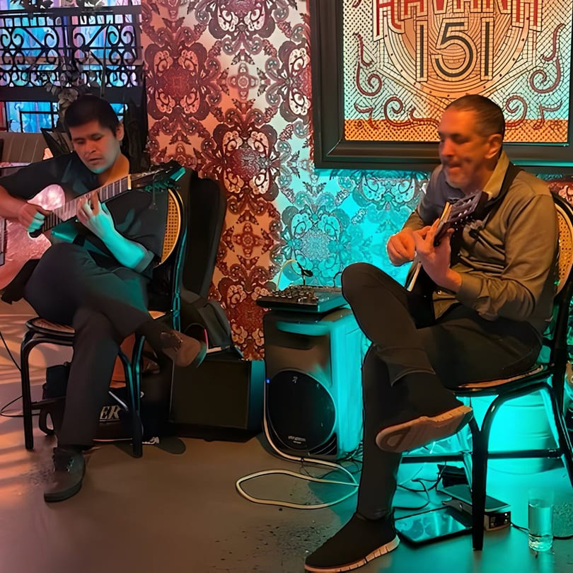 Two men playing guitars, illuminated by blue and orange light, against a patterned wall.