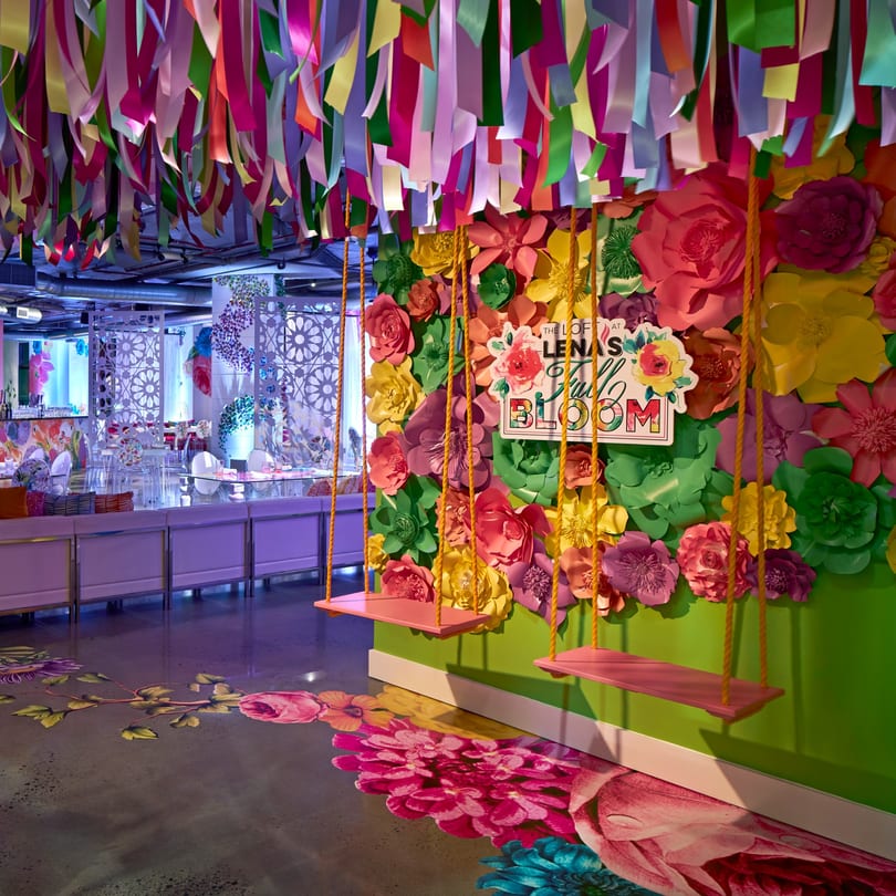 Colorful paper flowers and ribbons adorn a vibrant event space with two pink swings and floral floor decals.