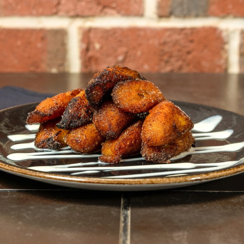 A plate of golden-brown fried Cuban plantains, drizzled with white sauce, against a brick wall.