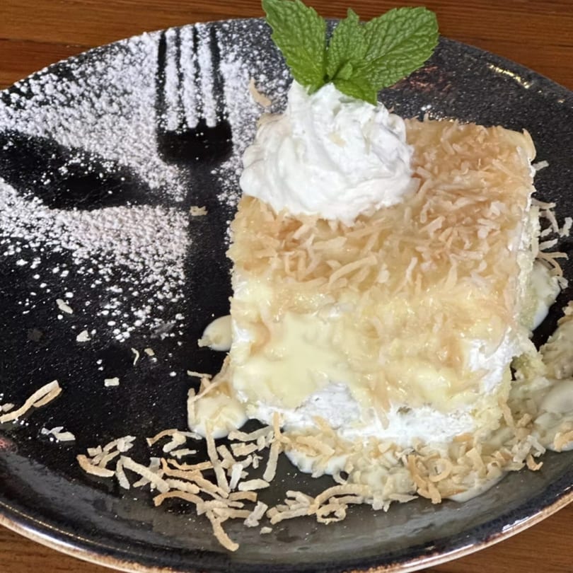 Coconut cake with whipped cream, mint, and powdered sugar on a dark plate.