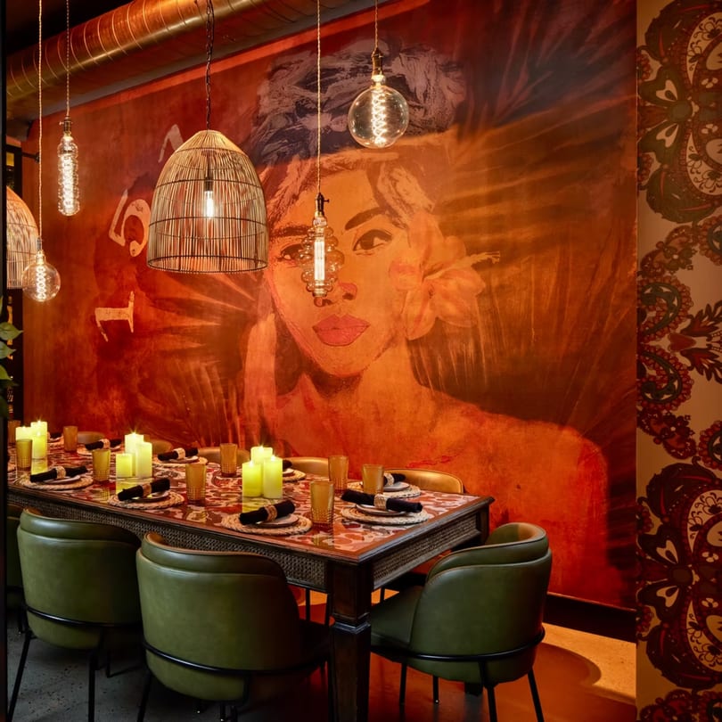 A dining table set with candles and green chairs, against a mural of a woman's face.