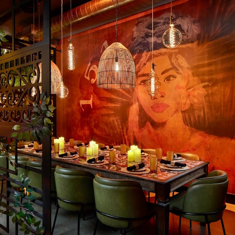 A long dining table set with candles and green chairs, against a mural of a woman's face.