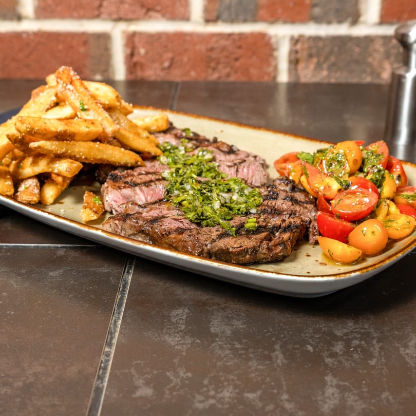 Grilled steak with chimichurri, a side of fries, and a tomato salad on a rectangular plate.