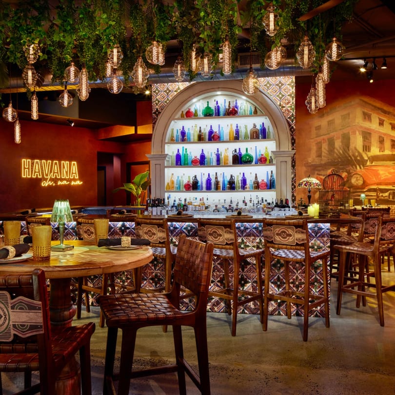Havana 151 restaurant interior with a neon sign, bar, and tables under hanging plants and lights.