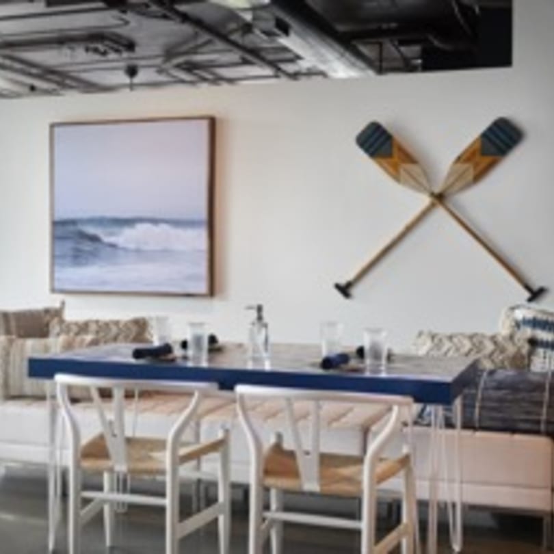 A dining area with a blue table, white chairs, a wave painting, and crossed oars on the wall.