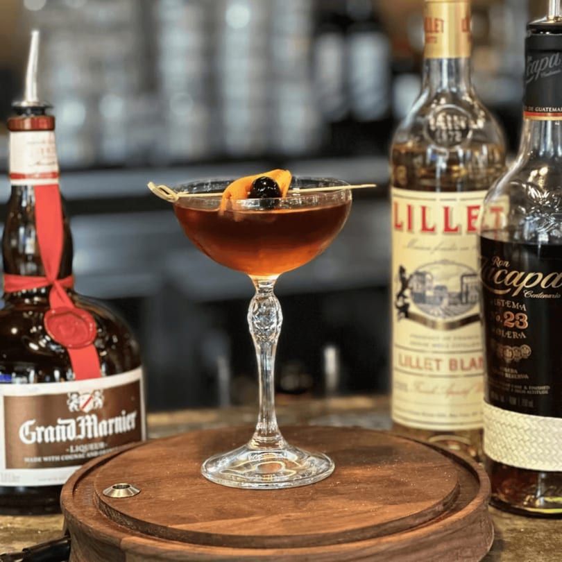 A Havana 151 cocktail, garnished with orange peel and cherry, sits on a wooden tray with liquor bottles.