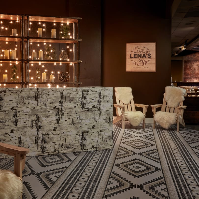 Cozy winter lodge decor with birch-covered bar, Adirondack chairs, string lights, and a patterned rug.