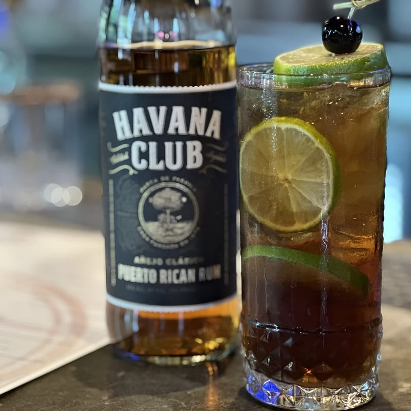 A Havana Club rum bottle next to a tall cocktail garnished with lime and a cherry.