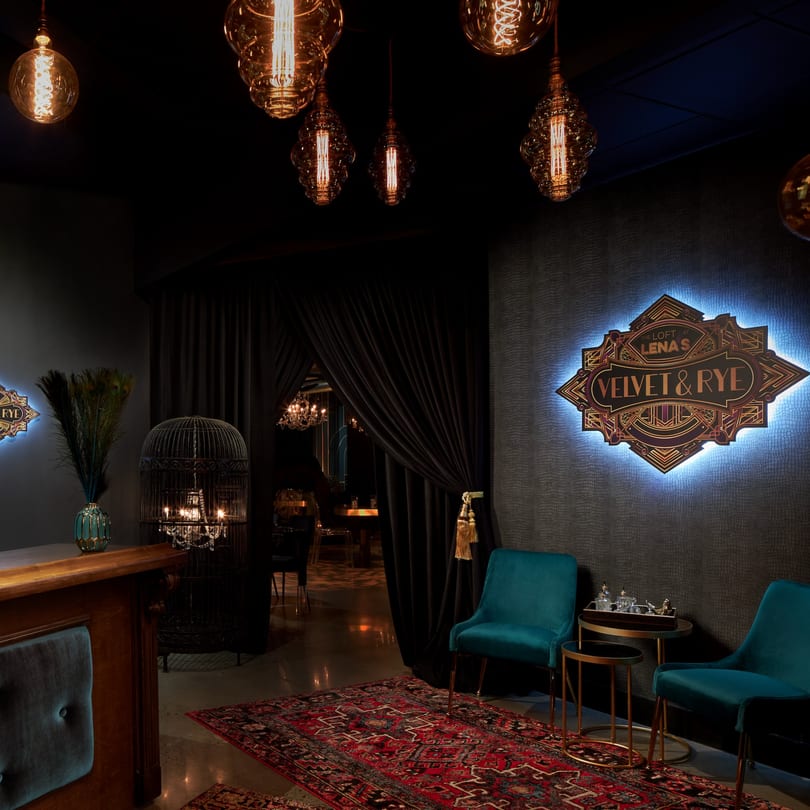 Dark, luxurious lounge entrance with velvet chairs, ornate rug, and glowing "Velvet & Rye" sign.