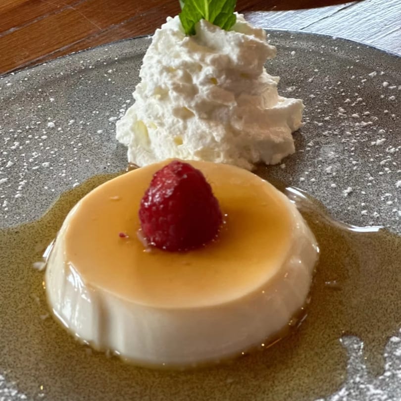 Cuban flan dessert with caramel sauce, a raspberry, whipped cream, and mint on a gray plate.