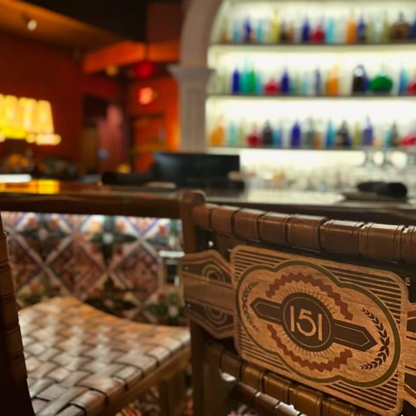 A Havana 151 bar stool with a decorative backrest and a blurred background of colorful bottles.