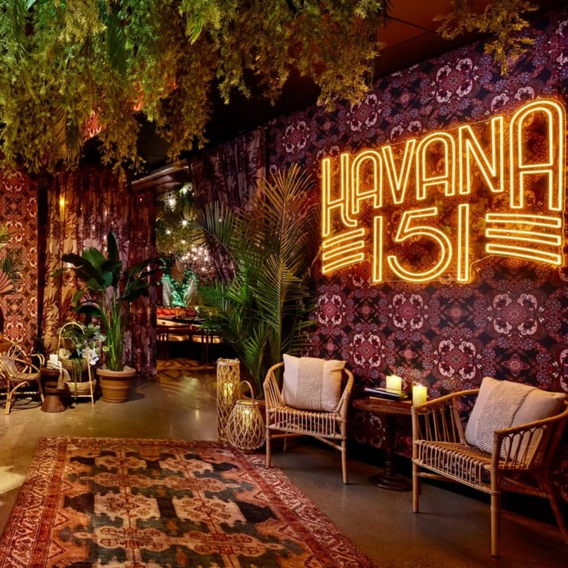 Havana 151 neon sign illuminates a lounge area with rattan chairs, plants, and patterned decor.