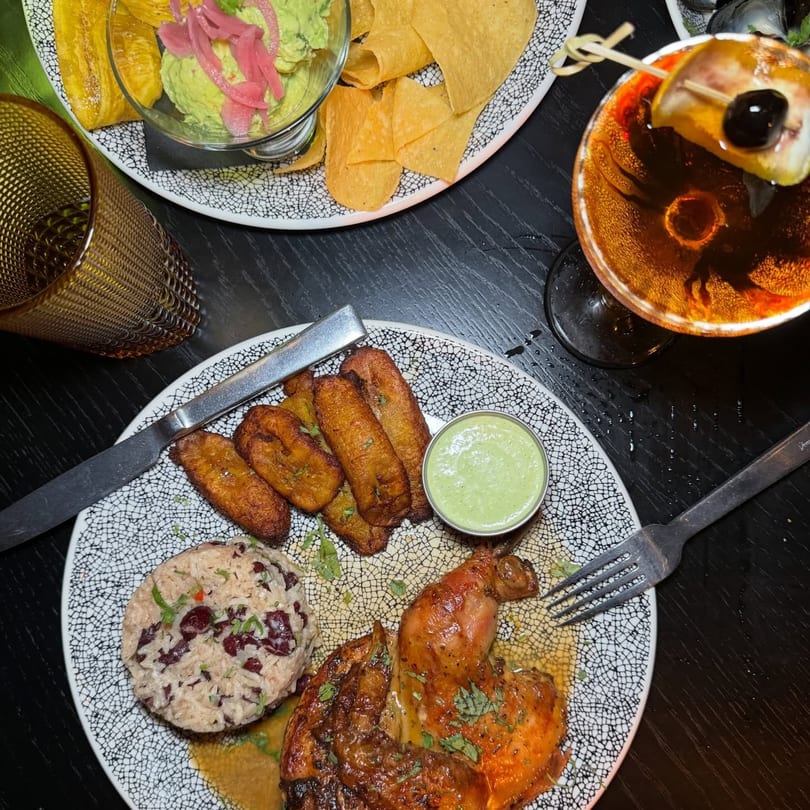 Cuban meal with roasted chicken, rice and beans, plantains, guacamole, chips, and a cocktail.