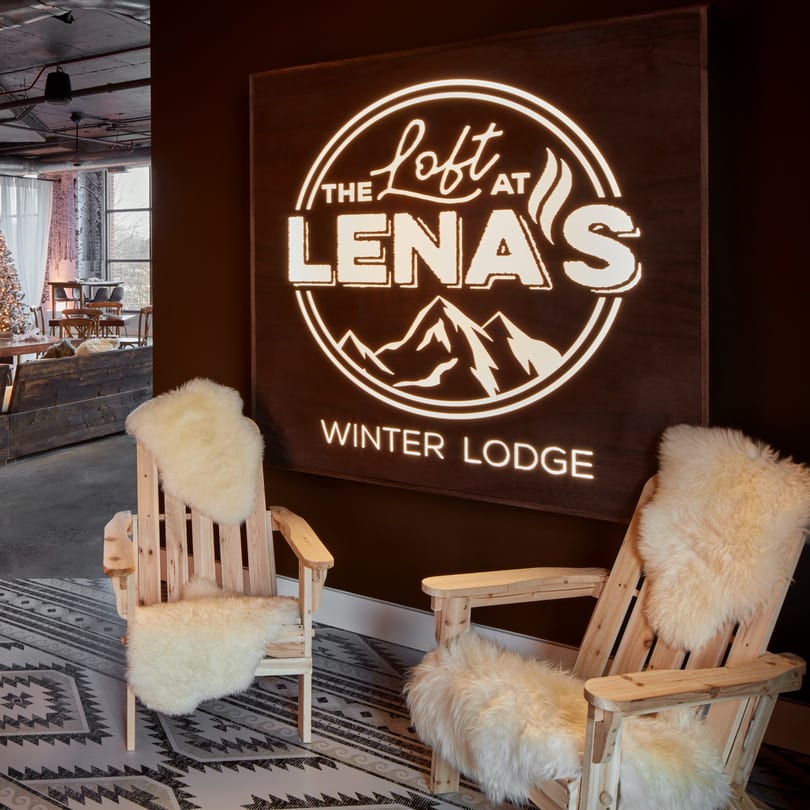 Two wooden Adirondack chairs with fluffy white throws sit before a glowing "The Loft at Lena's Winter Lodge" sign.
