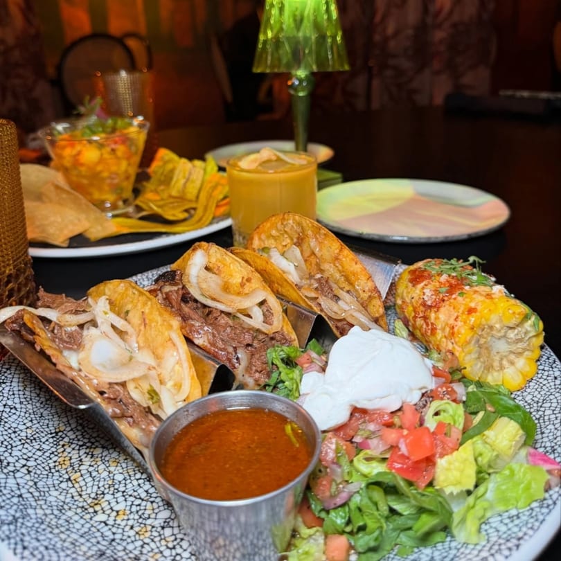 Cuban beef tacos with corn, salsa, and sour cream, alongside plantain chips and drinks.
