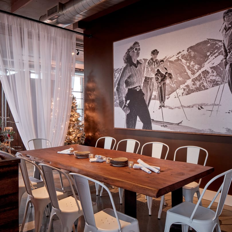 Long wooden dining table with white chairs, set against a large ski mural and sheer curtains.