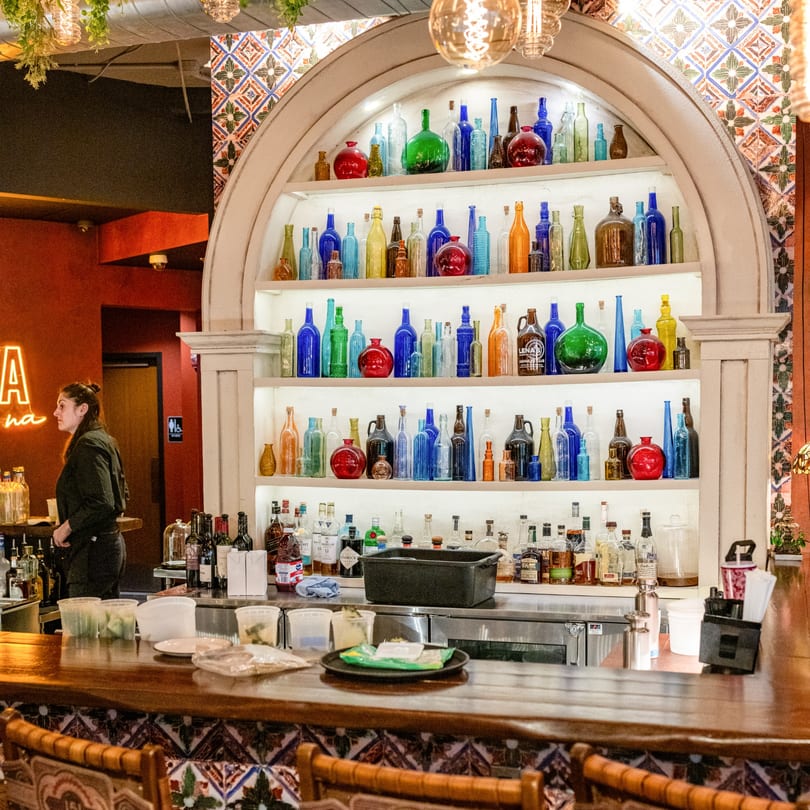 A bartender stands behind a wooden bar with colorful bottles displayed on shelves in a Cuban restaurant.