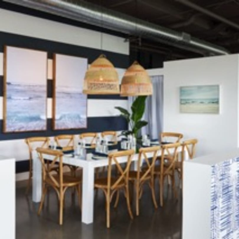 A long white dining table with wooden chairs, wicker lamps, and ocean art on a striped wall.