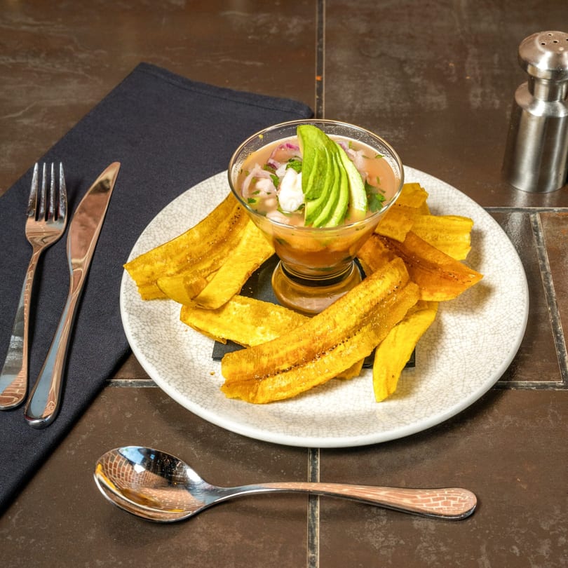 Cuban ceviche with avocado and plantain chips on a white plate with silverware and a salt shaker.