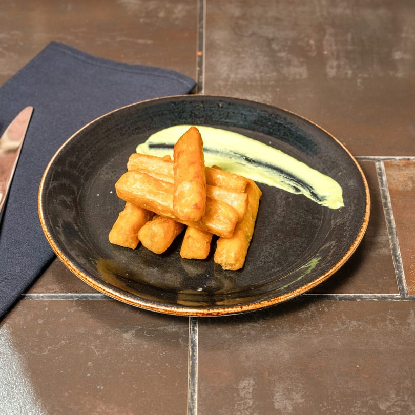 Golden-brown yuca fries with a swirl of light green sauce on a dark plate, next to a napkin and knife.