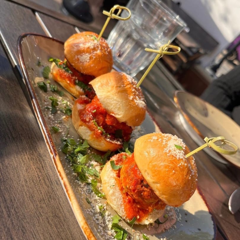 Three Cuban meatball sliders on a platter, garnished with herbs and cheese, on a wooden table.