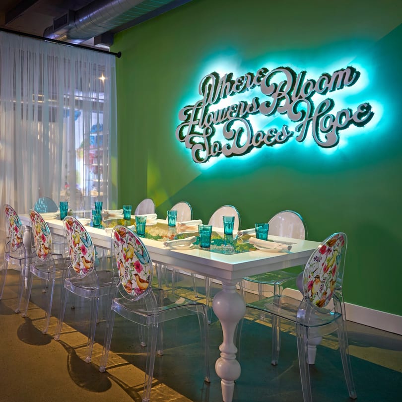 A long white dining table with clear chairs and teal glasses, against a green wall with a neon sign.