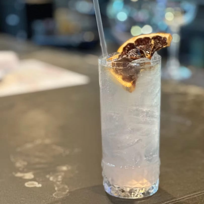 A tall glass of clear, iced cocktail with a dried orange slice and straw on a bar.