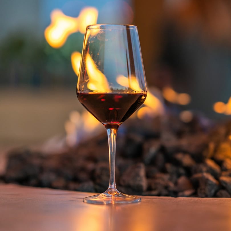 A glass of red wine sits on a table with a fire pit burning in the background.
