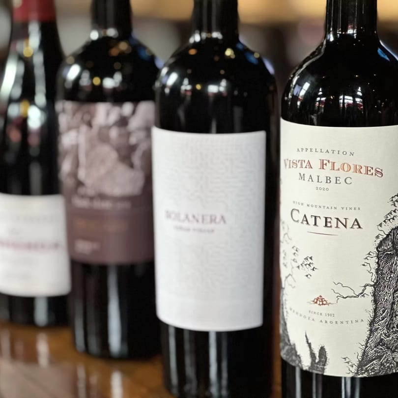 Four dark wine bottles with various labels, including "Catena Vista Flores Malbec," on a wooden surface.
