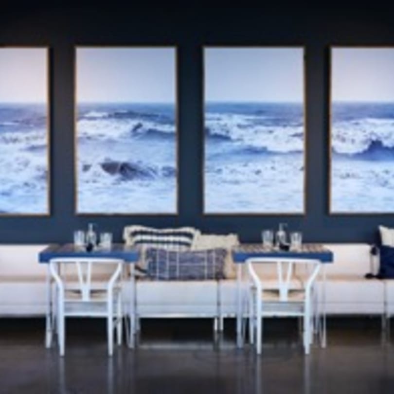Dining area with white banquette seating, blue tables, and large ocean wave art on a dark blue wall.