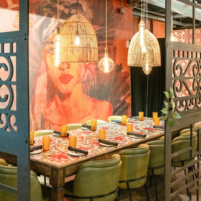 A long dining table with green chairs, amber glasses, and a vibrant mural of a woman in the background.