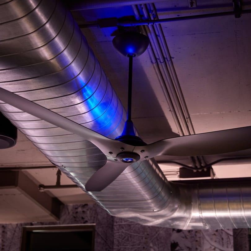 A modern ceiling fan with blue light hangs near exposed silver ductwork in a dimly lit room.