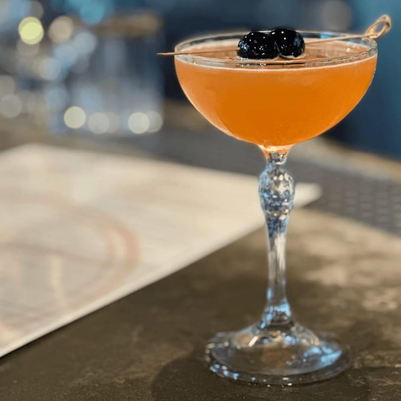 A vibrant orange cocktail in a coupe glass, garnished with two dark cherries on a pick.