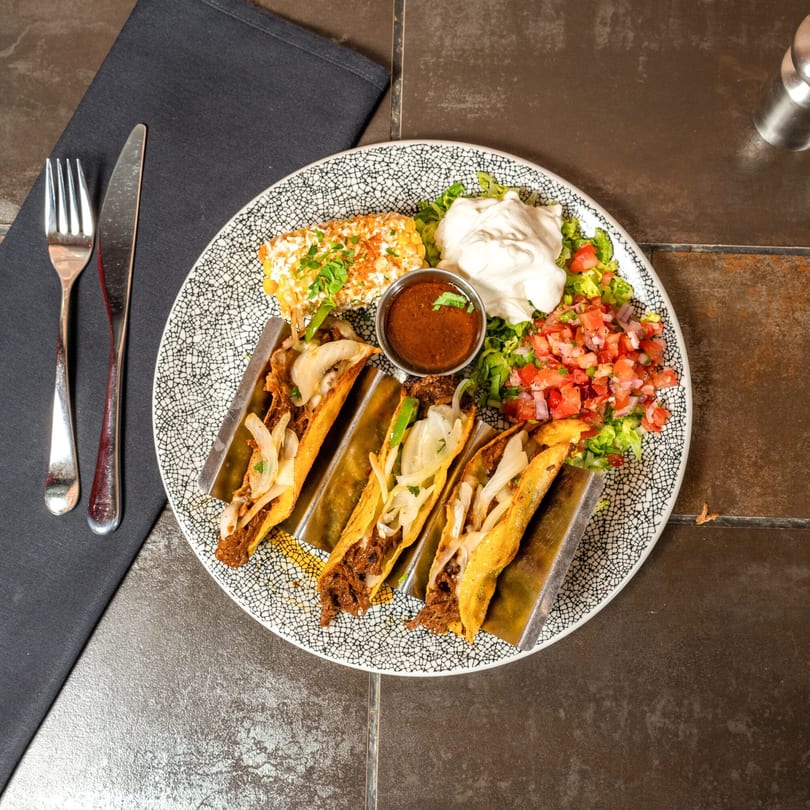 Four Havana 151 tacos with elote, salsa, and sour cream on a patterned plate.