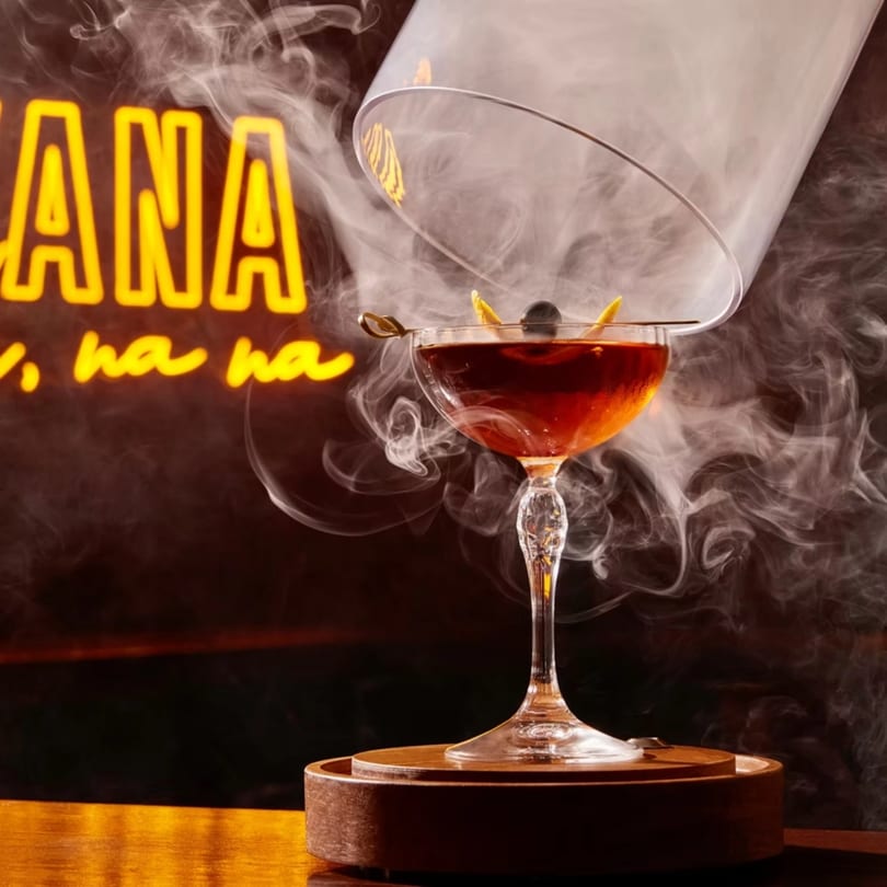 Smoked cocktail with a cherry and orange peel garnish, with a neon "HAVANA" sign in the background.