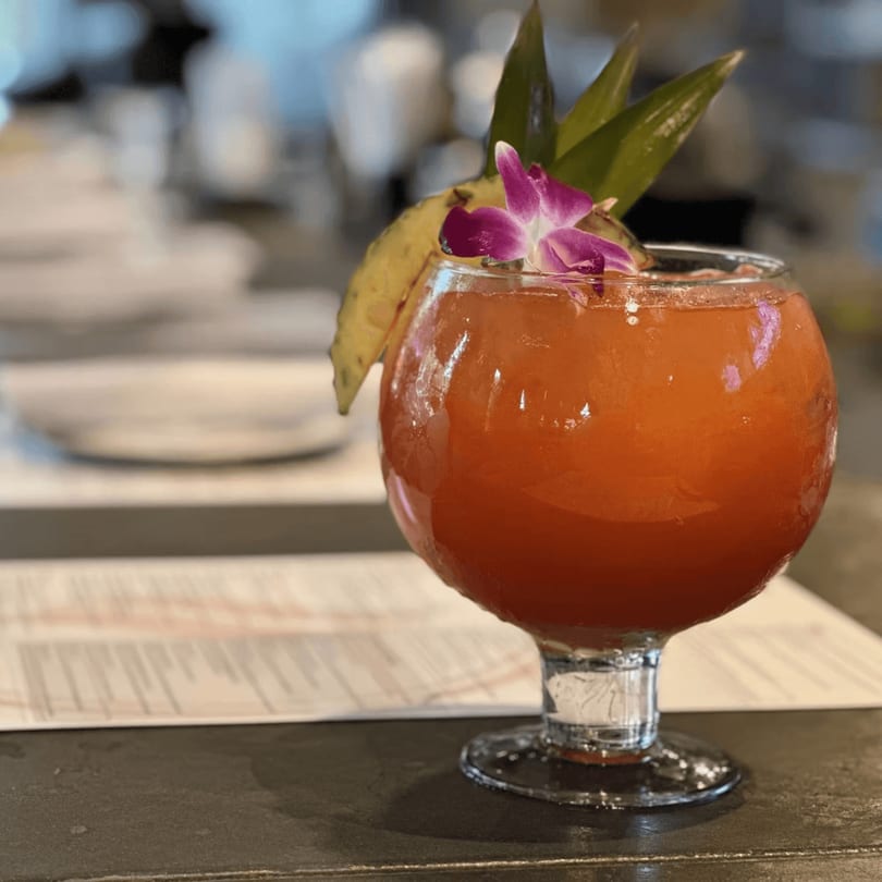 A vibrant red Cuban cocktail, garnished with pineapple and an orchid, sits on a dark bar.