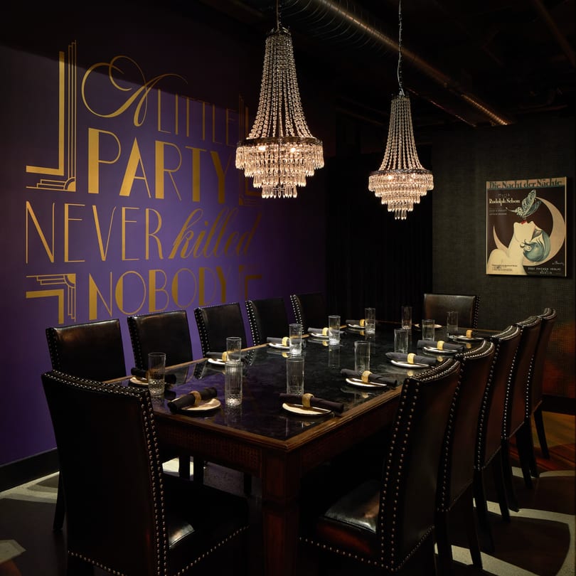 A private dining room with a long table, leather chairs, chandeliers, and a purple wall with gold text.