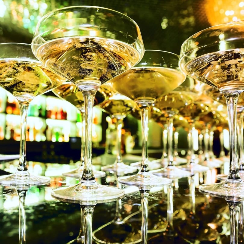 Row of champagne coupes filled with sparkling golden liquid, reflecting a vibrant bar background.